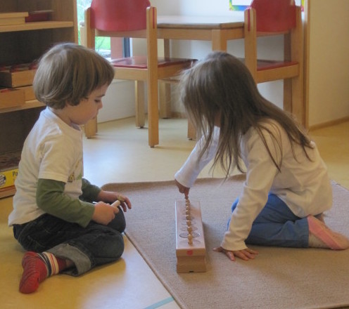 Zwei Kinder sitzen auf einem Teppich und arbeiten gemeinsam mit einem Einsatzzylinder-Block aus Holz