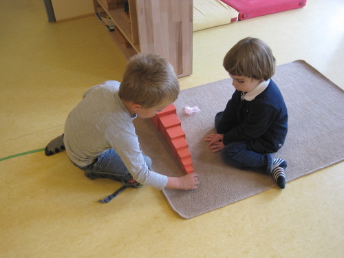 Zwei Kinder bauen auf einem Teppich gemeinsam mit der roten Treppe aus Holz