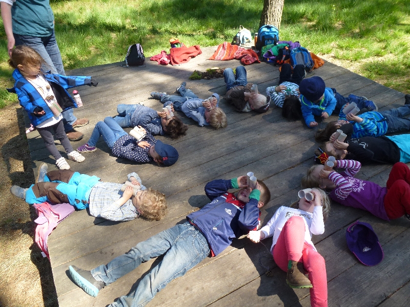 Kinder liegen auf einer Holzplattform im Grünen und schauen in den Himmel