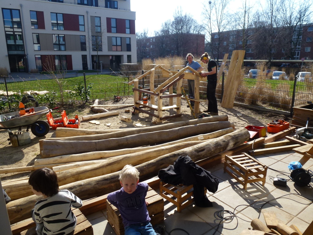 Handwerker bauen das Gerüst des Spielhauses auf, Kinder schauen im Vordergrund zu