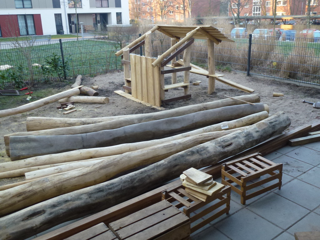Das Holzgerüst des Spielhauses mit Dachbalken steht, davor liegen Baumstämme und Holzkisten